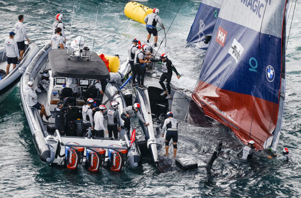 A half-sunken America's Cup boat assisted by a powerboat and crewmembers using floats to keep the boat from fully sinking.