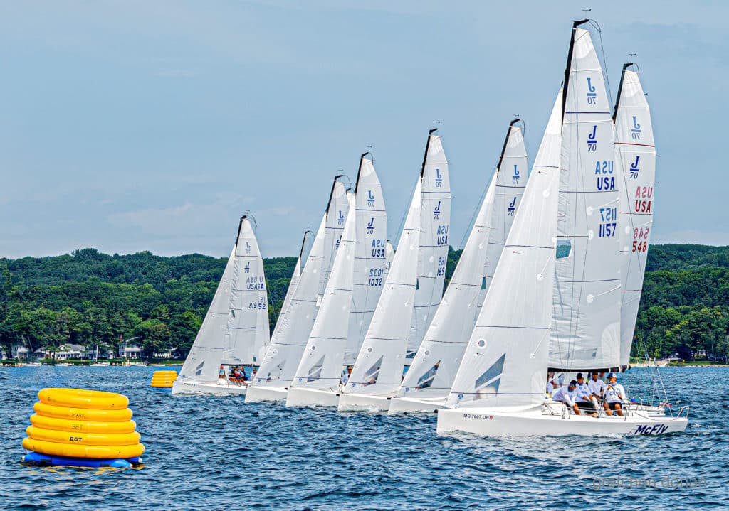 J/70 Sailboats lining up to start on a blue sky summer day with a yellow buoy in the foreground.