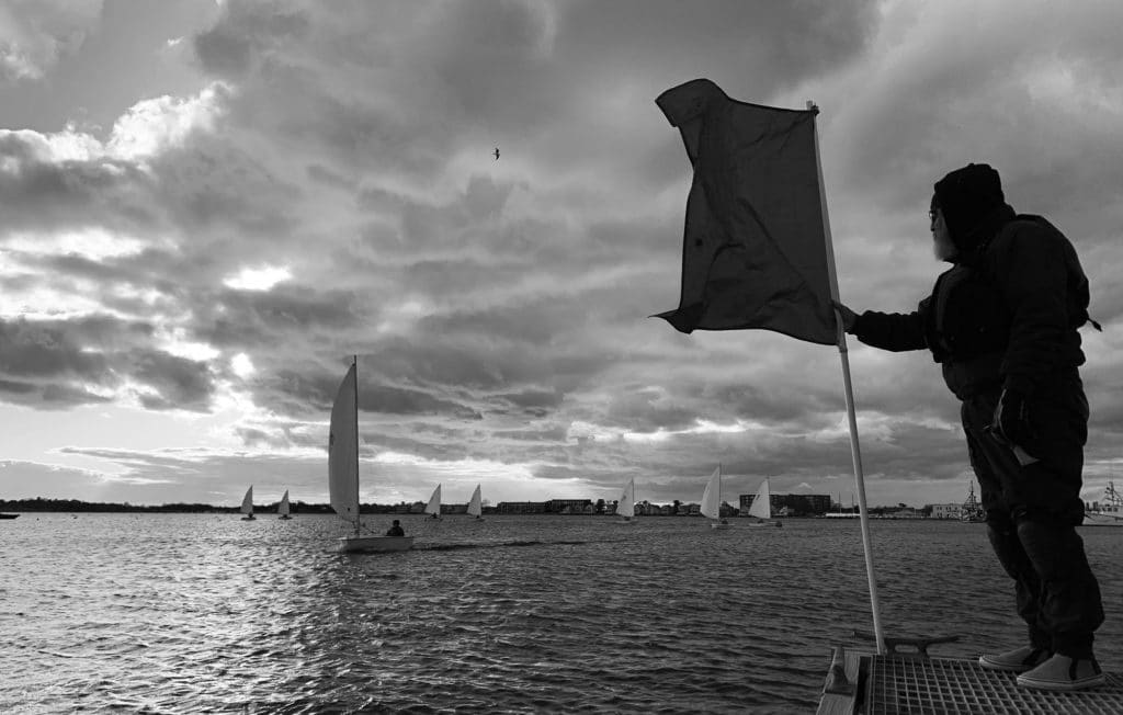 Man standing on dock with a flag to finish a sailboat racer off in the distance.