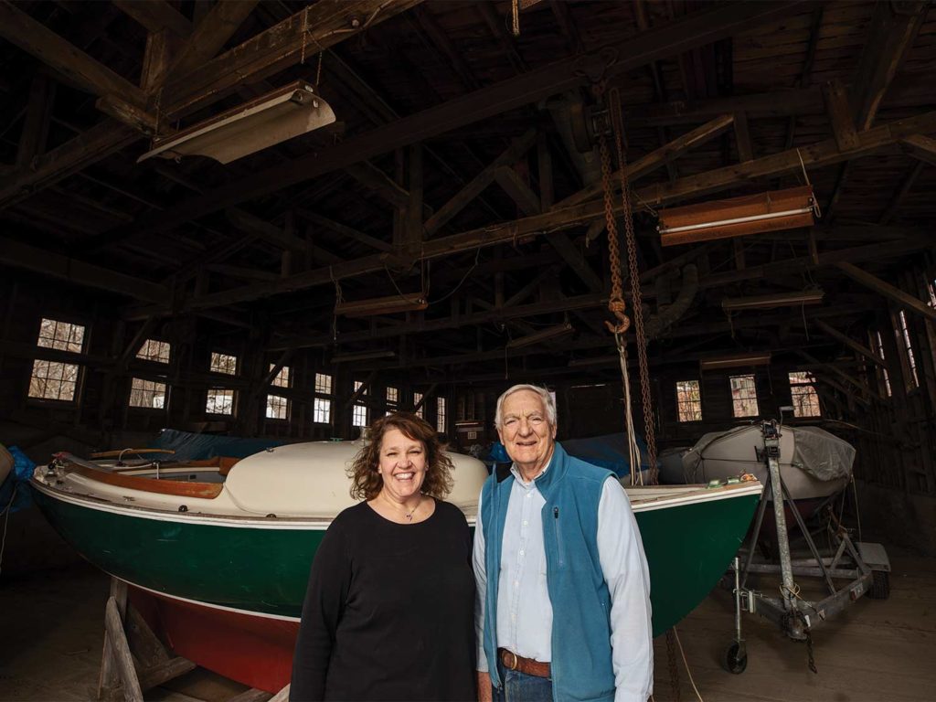 A man and woman stand in front of a sailboat hull.
