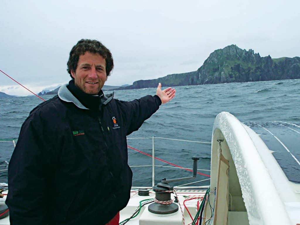 A man stands on a sailboat and gestures to an island in the distance.