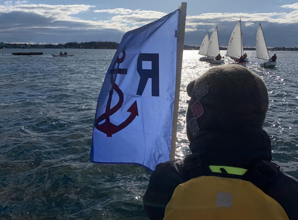 Vantage point of a sailboat race start from the committee boat with flag and fleet of boats.
