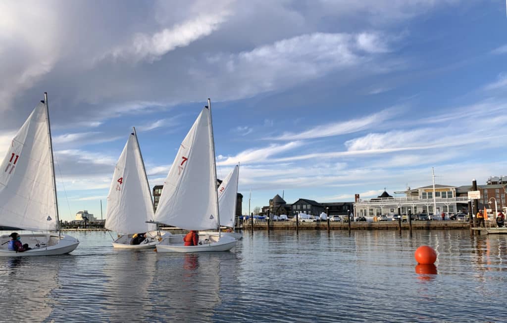 Small sailboats on a glassy harbor approaching and orang buoy that marks the finish line.