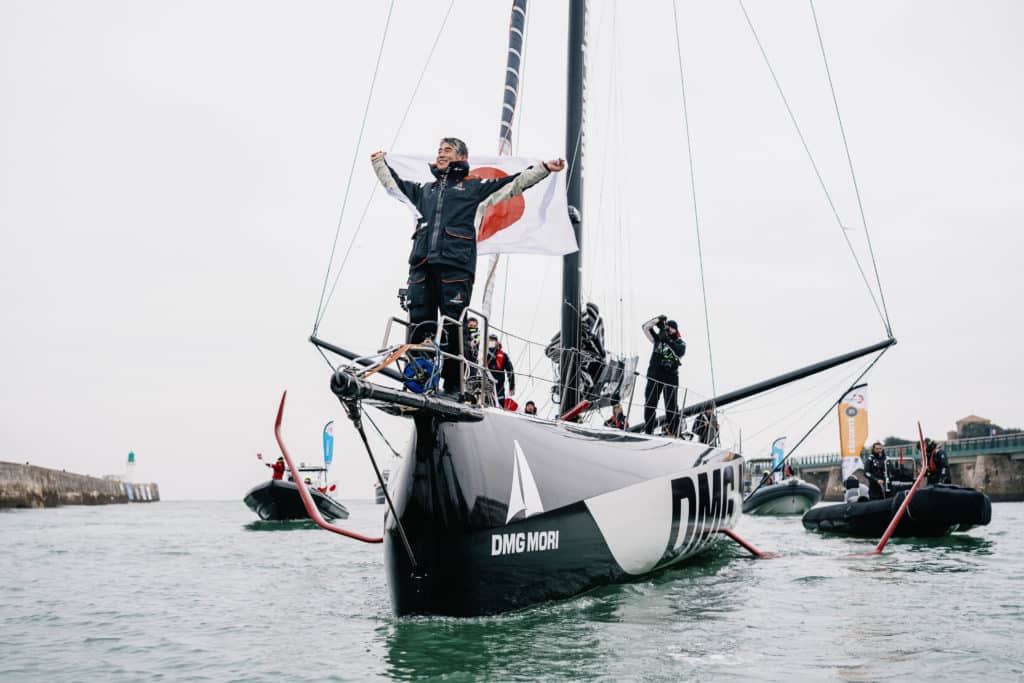Vendee Globe solo skipper standing at the bow of his boat with a Japanese flag, celebrating his finish.
