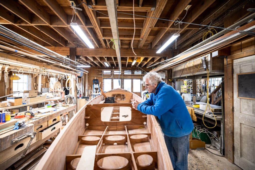a man stands over a wooden kit-build sailboat in his woodshop and the viewer can see the inner framing of the vessel