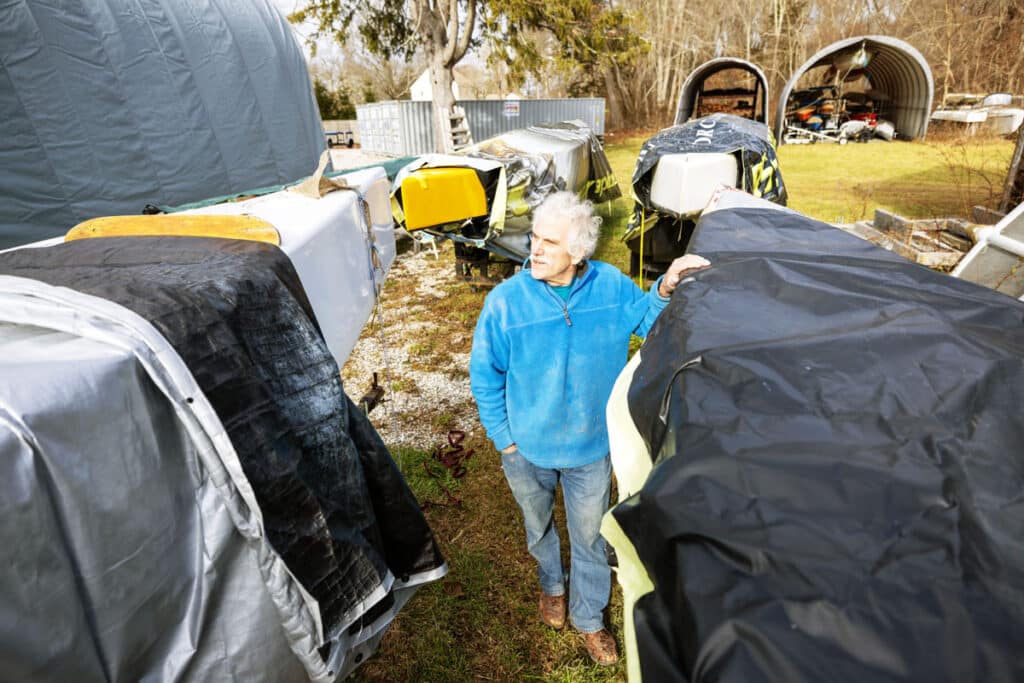 A man walls through an arrangement of stored vessels, boat hulls covered in tarps.