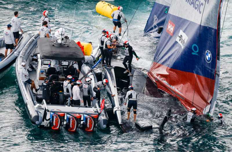 A half-sunken America's Cup boat assisted by a powerboat and crewmembers using floats to keep the boat from fully sinking.