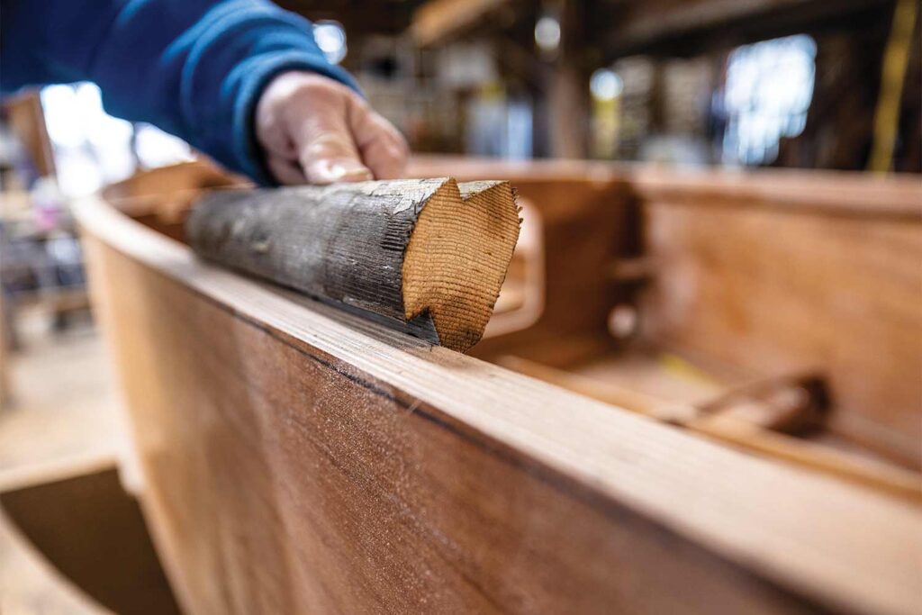 A man holds a length of wood up next to a worked, shaped, hull of a sailboat.