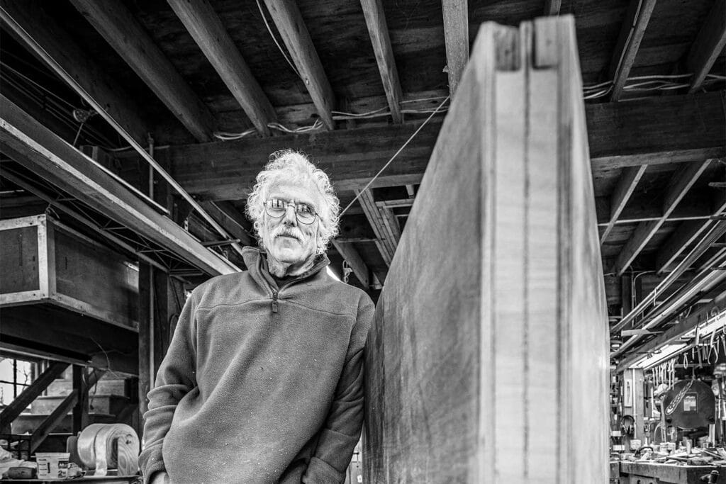 A black and white image of a man standing next to his sailboat build in progress.