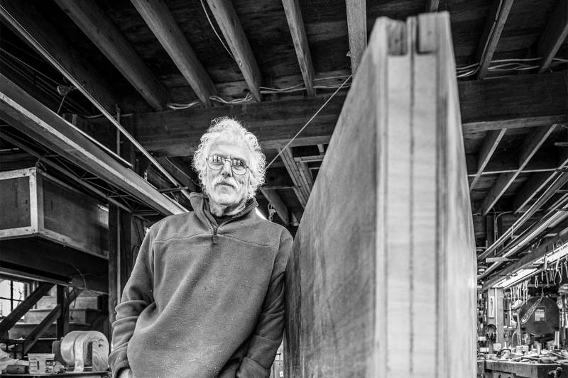 A black and white image of a man standing next to his sailboat build in progress.